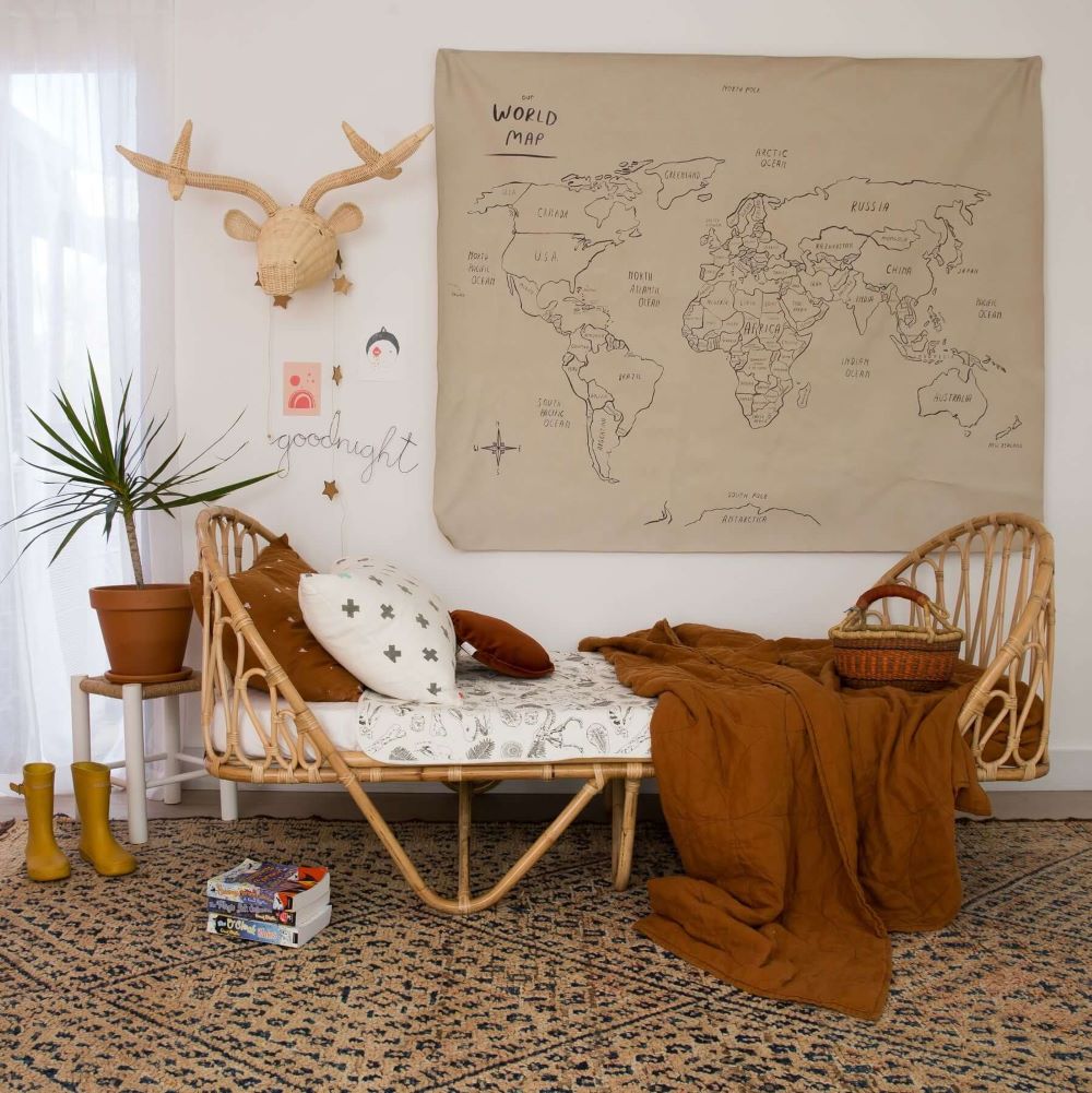 Cozy children’s bedroom with rattan bed, dinosaur print bedding, brown quilt, world map wall hanging, wicker deer head decor, indoor plant, and patterned rug.